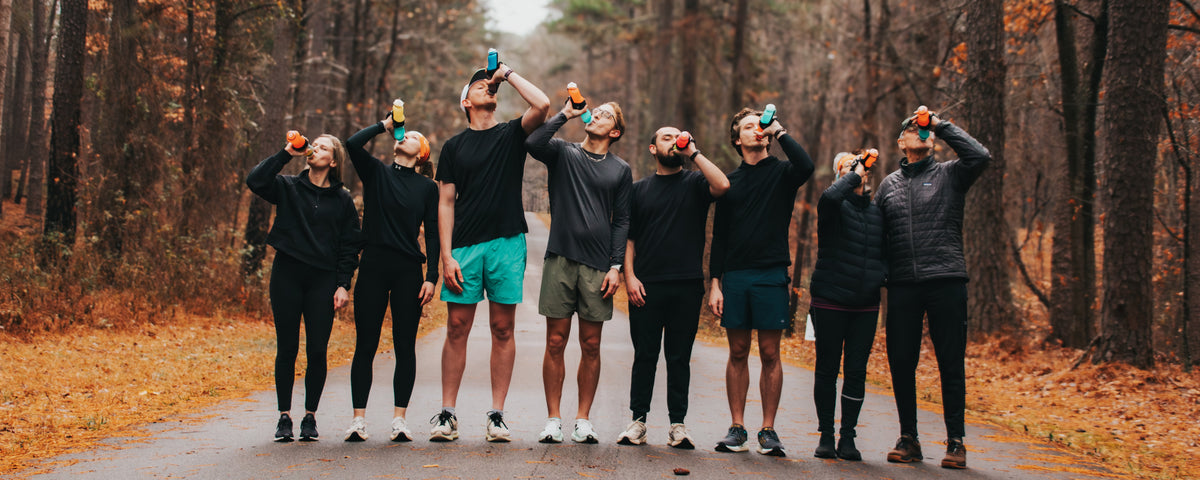 A group of eight people standing on a road in a wooded area, drinking from small bottles held up to their mouths. They are dressed in dark athletic clothing, with autumn trees and fallen leaves in the background.